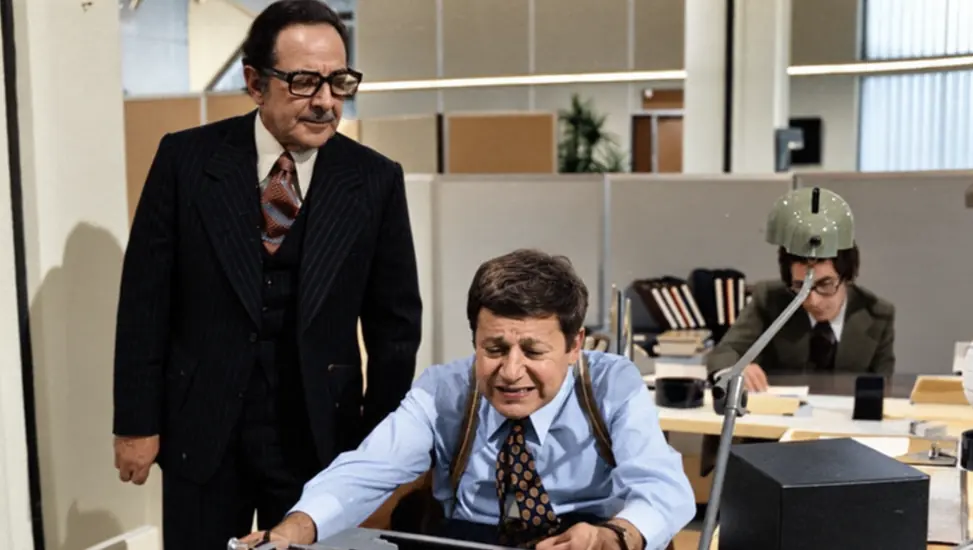 Fantozzi at his desk under his superior’s gaze, in an office.